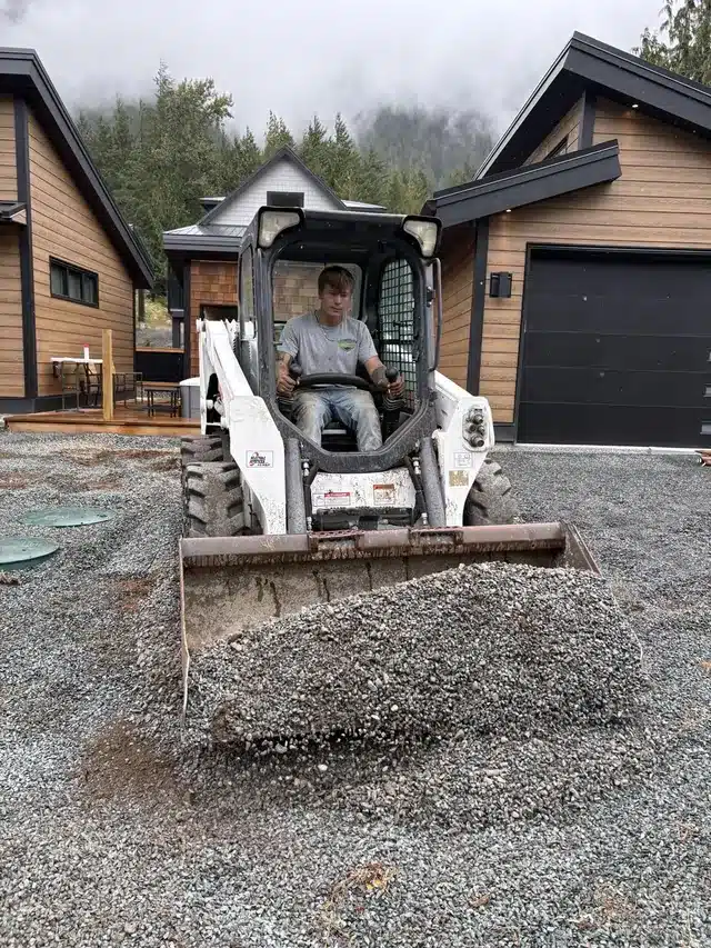 Skid steer moving a bucket of gravel on a new Fraser Valley home site, the kind of clean stone used in drainage trenches