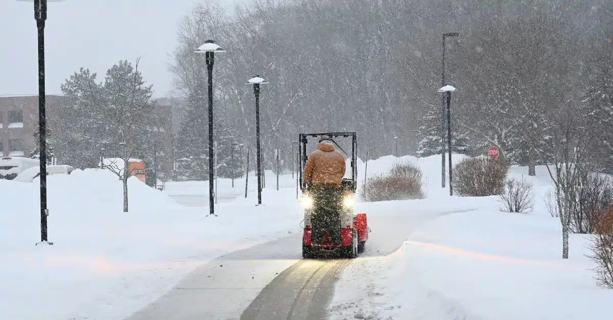 Compact sidewalk plow clearing a snow-covered walkway past lamp posts, the kind of path a strata snow removal contract in Chilliwack should cover